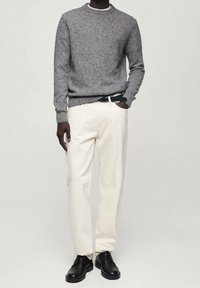 Homme portant un pull gris moucheté, un pantalon écru avec une ceinture noire, et des chaussures habillées en cuir noir, debout devant un fond uni.