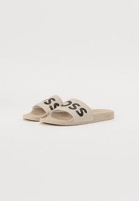Beige Riemchensandalen mit einem breiten Riemen, der das schwarze "BOSS"-Logo trägt. Glatte Textur und flache Sohle. Minimalistisches Design mit offener Zehe.