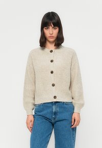 Kvinne med mørkt hår som har på seg en beige knappet strikkegenser og høy-hofte blå jeans, står mot en ensfarget hvit bakgrunn.