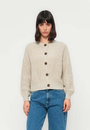 Kvinne med mørkt hår som har på seg en beige knappet strikkegenser og høy-hofte blå jeans, står mot en ensfarget hvit bakgrunn.