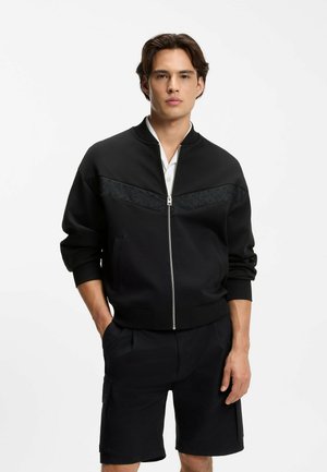 Jeune homme portant une veste noire zippée sur une chemise blanche et un short cargo noir, debout avec une main dans la poche sur fond blanc.