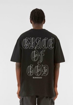 Osoba z krótkimi dredami nosząca czarny t-shirt z dużym białym gotyckim napisem "GRACE OF GOD" na plecach.