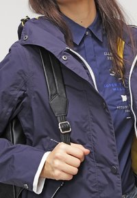 Navy blue waterproof jacket with a hood, zip front, and snap closures. Features yellow interior lining and contrasting white cuffs. Black backpack strap visible.