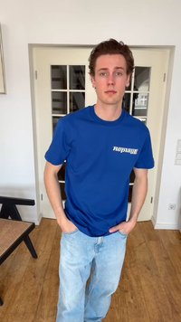 Jeune homme aux cheveux bruns debout à l'intérieur, portant un t-shirt bleu « Replay » et un jean bleu clair, les mains dans les poches, sol en bois et porte en verre derrière.