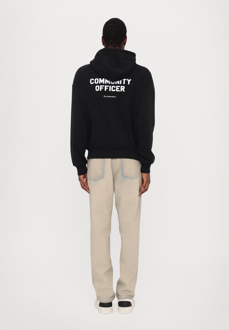 Black hoodie with "COMMUNITY OFFICER" text on the back, paired with light beige pants, featuring side pockets and a relaxed fit.