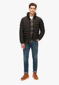 Giacca a vento nera con zip frontale e colletto alto, indossata sopra una camicia di flanella a quadri. Abbinata a jeans blu e stivali in pelle marrone con lacci.