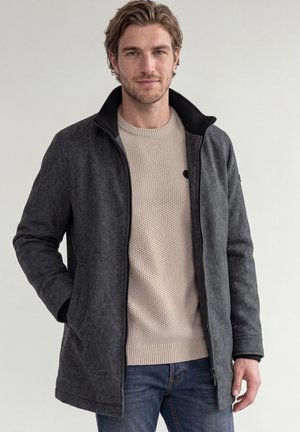 Homme aux cheveux châtain clair portant une veste grise zippée sur un pull en maille beige et un jean bleu, debout, une main dans la poche de la veste.