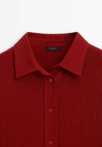 Chemise rouge texturée avec un col, présentant une patte de boutonnage et un subtil détail de côtes. Fabriquée en tissu léger, idéale pour un style décontracté.