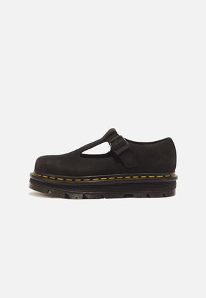Chaussure Mary Jane en cuir noir à semelle épaisse nervurée et coutures jaunes, avec bride en T et fermeture à boucle sur fond uni.