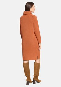 Femme de dos portant une robe-pull longue couleur rouille avec un col haut et des bottes en daim marron montant jusqu'aux genoux.