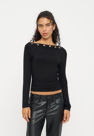 Jeune femme aux cheveux bouclés porte un haut noir à manches longues avec des œillets métalliques au niveau de l'encolure et un pantalon en cuir noir sur fond blanc.