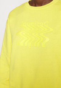 Sudadera amarilla con cuello redondo acanalado y un logo ondulado ligeramente elevado del mismo color. Tela de algodón suave y con textura.