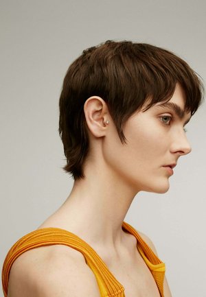 Pelo corto castaño, vistiendo una camiseta sin mangas de color naranja con textura acanalada. Perfil lateral mostrando un pendiente y una estructura facial suave.