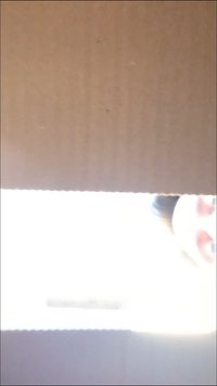 Cardboard box partially visible, revealing a bright light and a blurred face with dark hair. Box texture appears smooth, light brown.