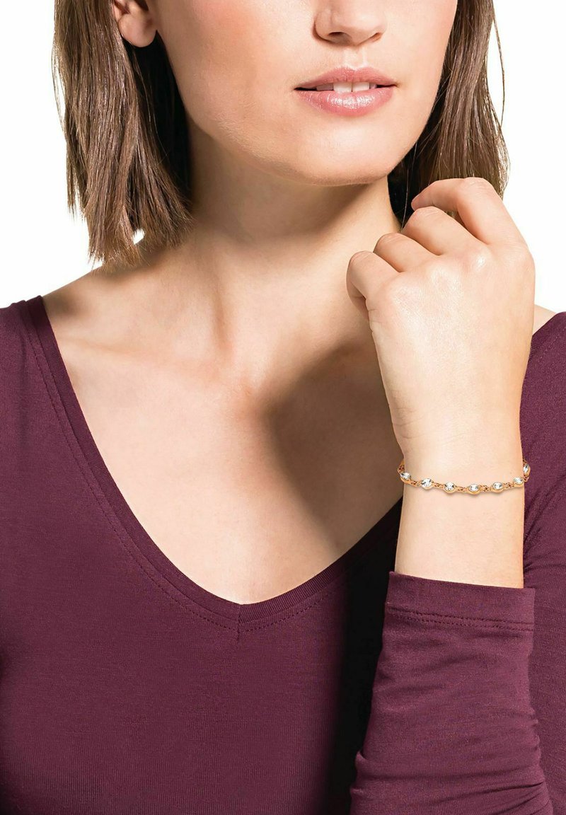 Pulsera de tono dorado con eslabones ovalados y piedras transparentes, llevada en una muñeca. El fondo es neutral, complementando la parte superior de burdeos oscuro.