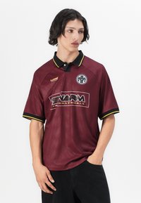 Πολύχρωμο πόλο μπλουζάκι σε μπορντό χρώμα με μαύρο γιακά, κίτρινες λεπτομέρειες, υφασμάτινη υφή και λογότυπο. Διαθέτει κοντά μανίκια και άνετη εφαρμογή.
