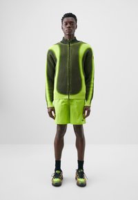 Tume rohelise ja erksate neoonroheliste detailidega kootud tõmbluku jakk; kombineeritud põlvekohaliste neoonlühikeste pükstega. Tekstuuriga kangas ja mustad sallid.