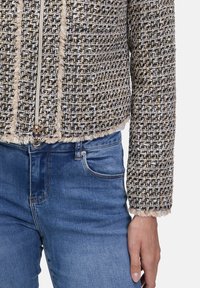 Tkana kurtka w czerni, bieli i złocie, z zamkiem z przodu, postrzępionymi brzegami i długimi rękawami, zestawiona z niebieskimi dżinsami.