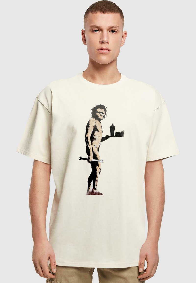 Beige oversize T-Shirt mit einem schwarz-braunen Grafikdesign eines Höhlenmenschen, der einen Knochen und ein Tablett mit Getränken und Pommes Frites hält.
