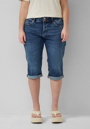 Personne portant un pantalon capri en jean bleu retroussé et des sandales blanches à plateforme, debout devant un fond gris uni.