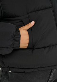 Hånd indsat i sidelomme på sort quiltet jakke med elastisk manchet, der viser en del af mørkegrå bukser nedenunder.