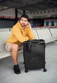 Černý tvrdý kufr s kulatým vzorem, s dvojitými koly a horní rukojetí, umístěný vedle sedící osoby na terminálu.