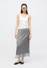 Grauer Satinsrock mit Spitzenabschluss, kombiniert mit einem weißen Tanktop. Das Modell trägt schwarze Ballerinas und weiße, transparente Strumpfhosen und steht vor einem schlichten Hintergrund.