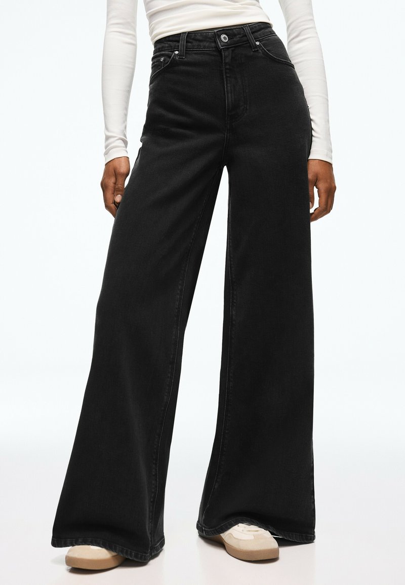 Personne portant un jean noir taille haute à jambes larges associé à des baskets beiges et un haut blanc ajusté à manches longues sur fond blanc.