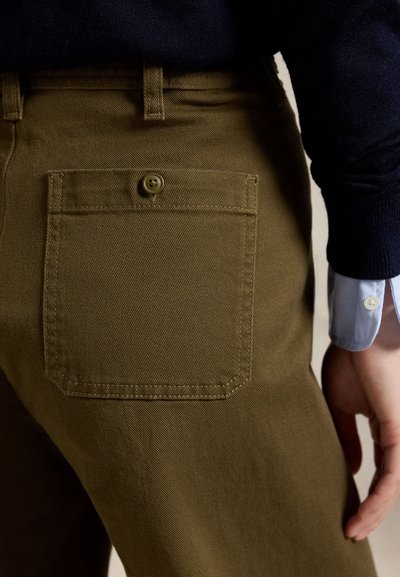 Nahaufnahme einer Person, die braune Hosen mit einer geknöpften Gesäßtasche und einen marineblauen Pullover über einer blauen Hemdmanschette trägt.