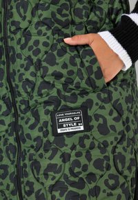Gilet trapuntato verde con stampa leopardata nera. Presenta una cerniera, tasca laterale e un'etichetta rettangolare con testo.