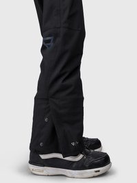 Underben med svarte snøbukser med trykknapper over svart og hvite Vans sneakers på en lys bakgrunn.