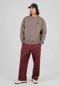 Brun genser med rund hals og trykket logo på brystet, kombinert med løstsittende burgunderbukser og beige sko. Avslappet stil.