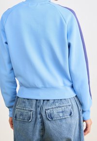 Felpa azzurra chiara con colletto e polsini a costine, caratterizzata da strisce laterali viola e un tessuto texture. Indossata con jeans a vita alta.
