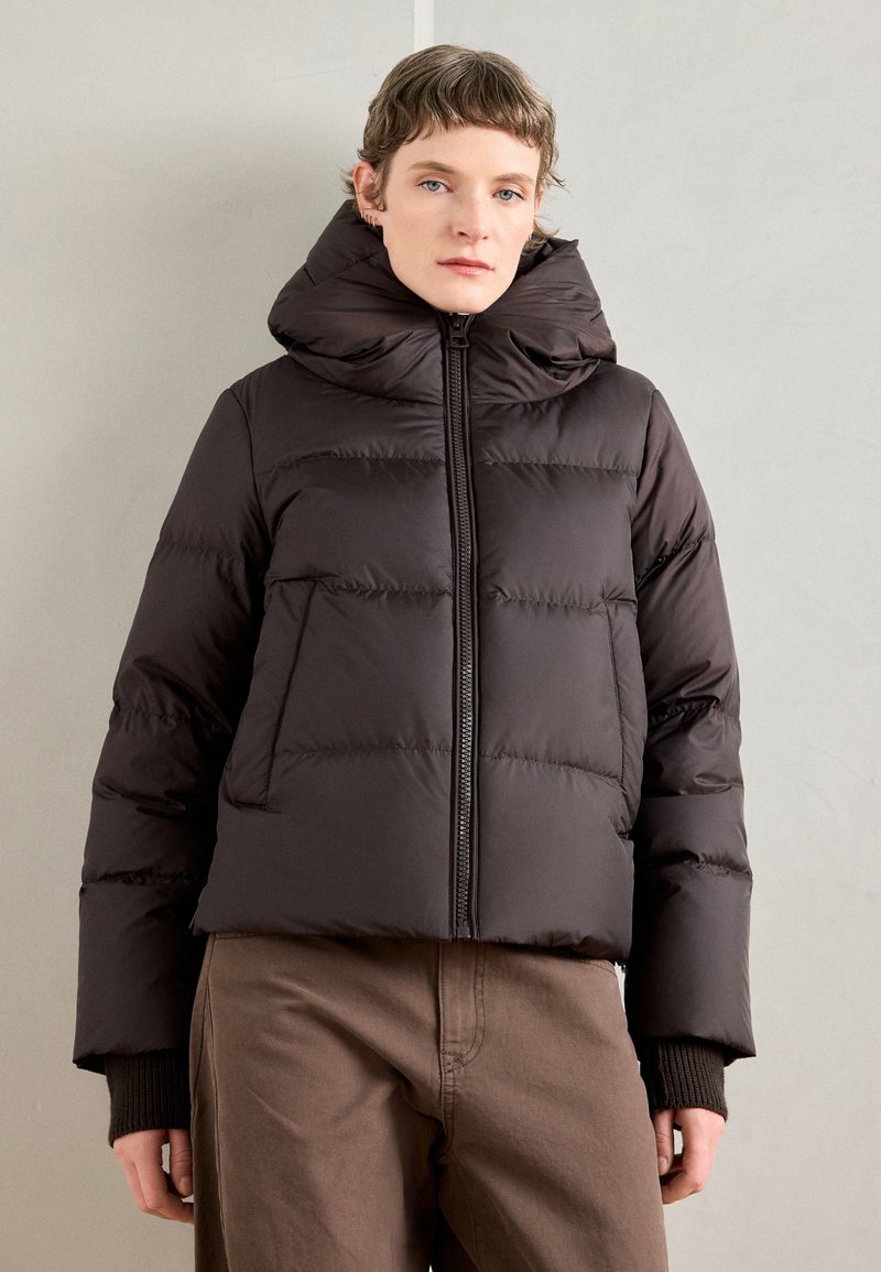 Personne portant une veste doudoune noire avec un col montant et un pantalon marron, se tenant contre un mur clair.
