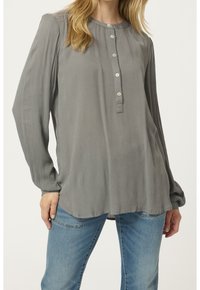 Blouse grise à manches longues avec une finition texturée, dotée d'un col rond et d'une patte de boutonnage à six boutons, associée à un jean bleu clair.