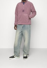 Ljuslila fleece-halv-zip-tröja med frampocket och logotypmärke, tillsammans med ljusblå baggy-jeans och svarta sneakers.