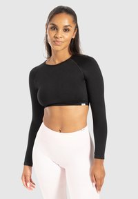 Zwart langzichtig crop top met een ronde halslijn en een gebandezoom. Het model draagt het met lichtroze hoge taille leggings. Glad textuur.