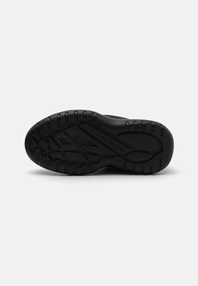 Semelle de basket noire avec une surface en caoutchouc texturé, présentant un motif de prise et un logo. Design courbé avec des éléments de traction surélevés.