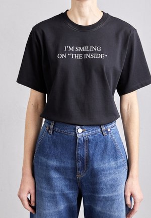 Person iført en sort T-shirt med hvid tekst "JEG SMILER INDENI", stoppet ned i højtaljede blå jeans.