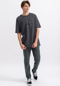 Grijze oversized T-shirt met een driehoekig grafisch ontwerp, gecombineerd met slim-fit groene broek en zwarte sneakers met witte veters.