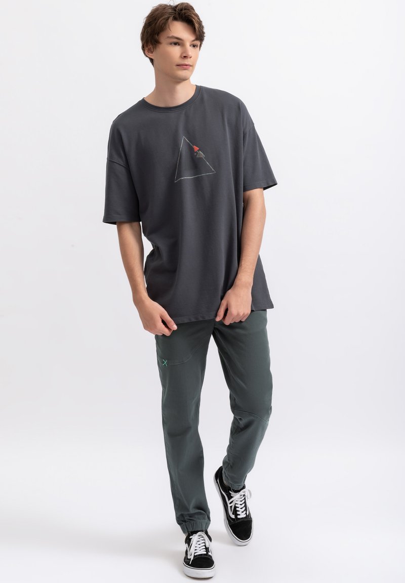 Grijze oversized T-shirt met een driehoekig grafisch ontwerp, gecombineerd met slim-fit groene broek en zwarte sneakers met witte veters.