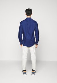 Mies seisoo kasvoin seinään ja hänellä on päällään sininen pitkähihainen paita, valkoiset housut ja moniväriset lenkkarit yksivärisellä taustalla.