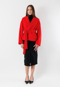 Cappotto avvolgente rosso con cintura e ampi revers, indossato sopra un dolcevita nero e una gonna a matita nera, abbinato a décolleté nere a punta.