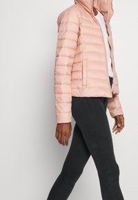 Chaqueta acolchada tipo puffer de color rosa claro, con capucha, cierre de cremallera y bolsillos laterales, combinada con jeans oscuros. Presenta una textura suave y un diseño ajustado.