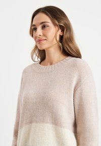Lichtroze trui met een ontspannen pasvorm, geribbelde halslijn en zachte textuur. Heeft een gradient van lichtroze naar crèmekleurig aan de onderkant.