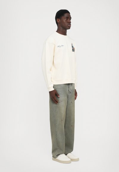 Jovem em pé de lado, vestindo um sweat-shirt creme com impressão gráfica, jeans cinzentos desbotados e soltos, e tênis brancos, em fundo liso.
