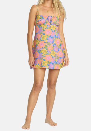 Femme portant une robe courte sans manches à fleurs orange, violettes et jaunes sur fond clair.