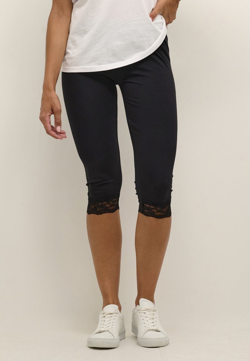 Femme portant des leggings noirs courts avec une bordure en dentelle, un t-shirt blanc et des baskets blanches, debout sur un fond uni.