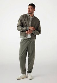 Kurtka bomberka w oliwkowej zieleni z prążkowanymi mankietami, zestawiona z luźnymi oliwkowymi spodniami i białymi sneakersami, charakteryzująca się gładką fakturą i minimalistycznym designem.