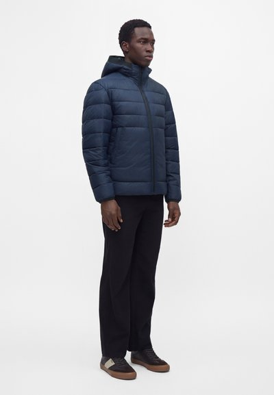Homme debout portant une veste matelassée à capuche bleu marine, un pantalon noir et des baskets marron et noires sur un fond blanc uni.
